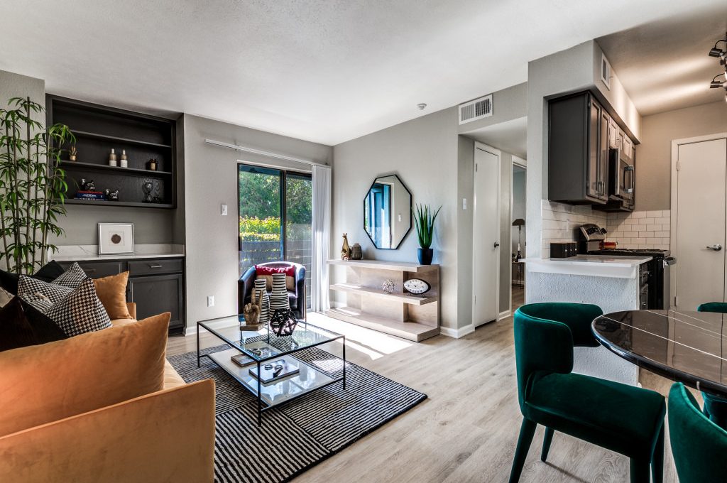 A cozy and modern living room features a comfortable orange sofa adorned with black and white checkered pillows. A glass coffee table sits atop a striped rug, adding a touch of elegance to the space. The room is illuminated by natural light streaming through a sliding glass door that opens to a balcony. A stylish dining area with green velvet chairs is adjacent to the kitchen, which boasts sleek black cabinets and stainless-steel appliances. The overall ambiance is inviting and perfect for both relaxation and socializing.